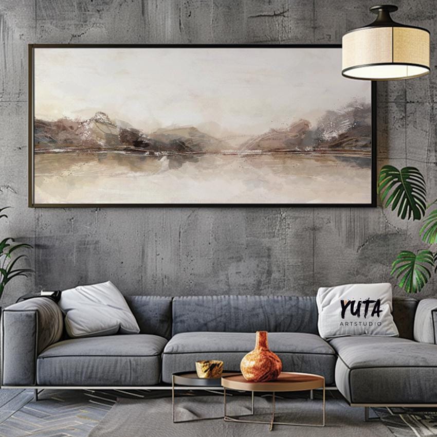 Framed neutral abstract landscape print above a grey sofa with books, plants, and ceramics.