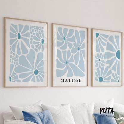 Three blue abstract botanical prints in light wood frames above a white sofa with books.
