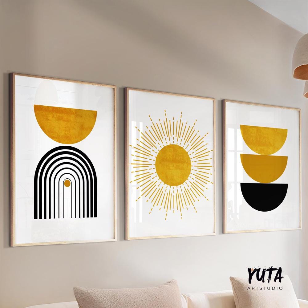 Three framed mustard yellow and black abstract prints displayed above a white sofa with books and a potted plant nearby.