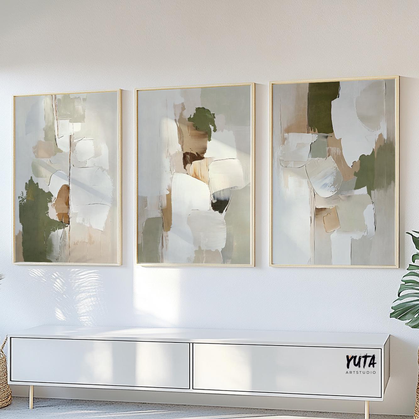 Three abstract olive green and beige prints in light wood frames above a white sideboard in sunlight