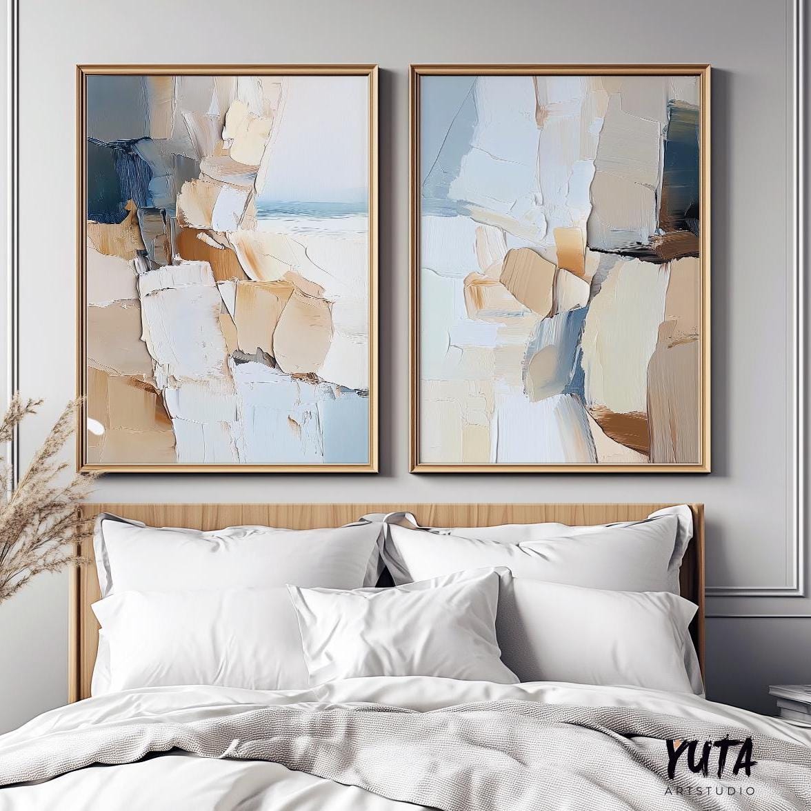 Two framed abstract coastal art prints in beige and blue displayed above a bed in natural light
